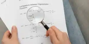 Tangan memegang kaca pembesar melihat rumus, menggambarkan proses menyusun dan menguji hipotesis penelitian.