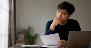 Pria Indonesia meninjau dokumen lamaran kerja di depan laptop, terlihat bingung karena belum mendapat panggilan.