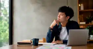 Pria Indonesia duduk di ruang kerja, memikirkan pilihan karier baru sambil melihat laptop dan catatan.