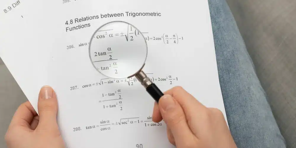 Tangan memegang kaca pembesar melihat rumus, menggambarkan proses menyusun dan menguji hipotesis penelitian.